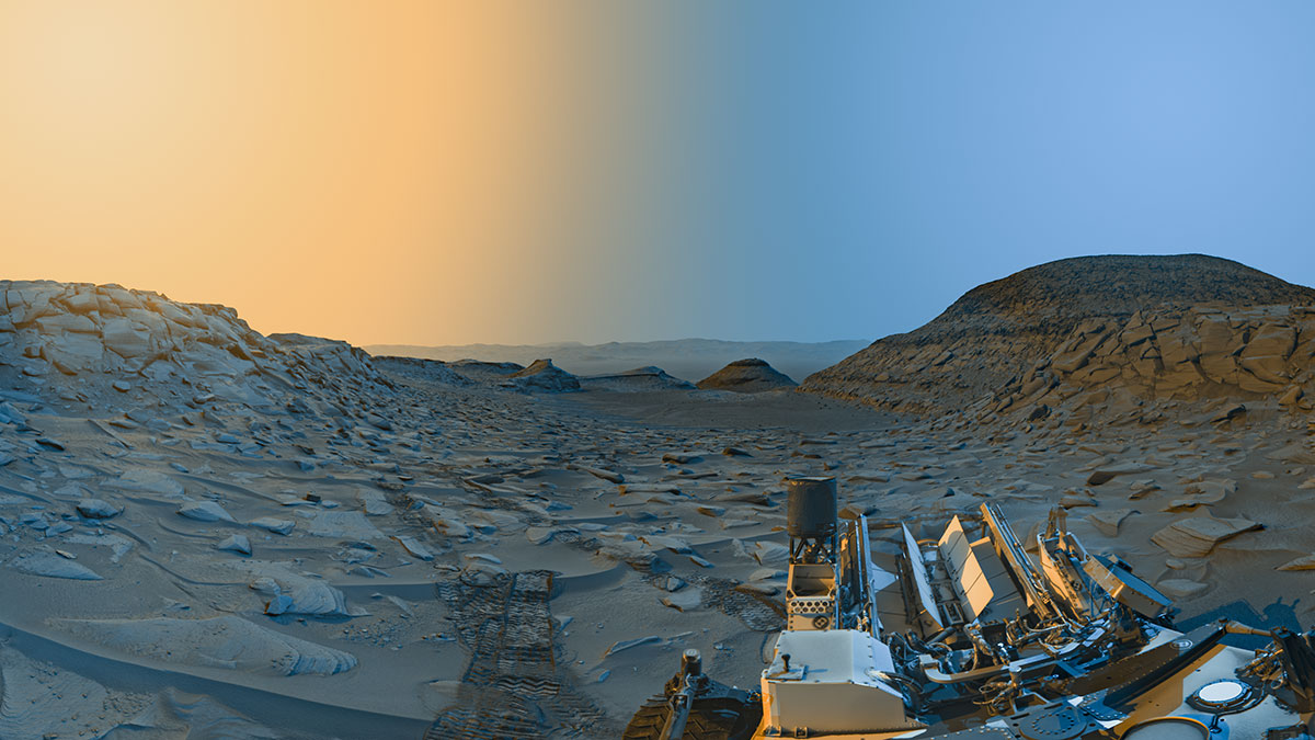 La foto coloreada del rover Curiosity de la NASA en Marte muestra una amplia vista de un valle rocoso en Marte. La mitad trasera del rover es visible en la parte inferior derecha, con las huellas de las ruedas en la superficie retrocediendo en la distancia. Esta vista estilizada tiene color añadido a un par de panorámicas en blanco y negro tomadas por el rover en la mañana y en la tarde. Los tonos azules y amarillos simulan la iluminación de la mañana y la tarde, respectivamente.